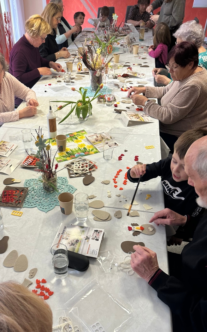 Auf dem Bild sieht man eine lange Tafel, an welcher unterschiedliche Menschen sitzen, sowohl Kinder als auch ältere Menschen. Alle sitzen zusammen und basteln. Auf dem Tisch stehen Blumen und es liegt viel Material zum Basteln durcheinander. 