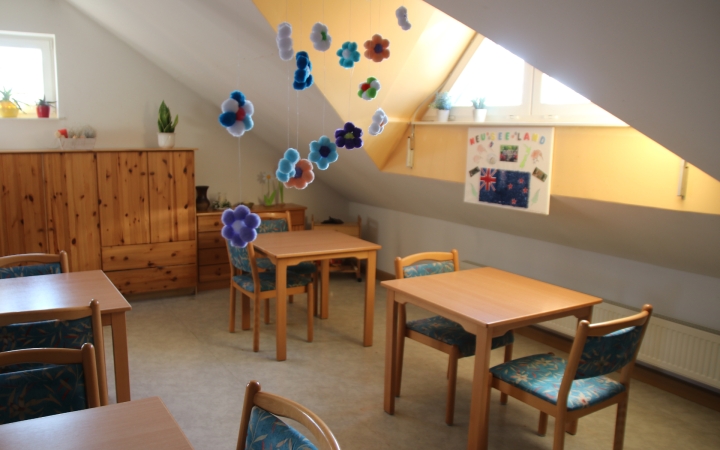 Heller Raum mit mehreren Holztischen und passenden Stühlen, die mit gemusterten Polstern bezogen sind. An der linken Wand steht ein großer Holzschrank, auf dem mehrere kleine Pflanzen und Dekorationsgegenstände platziert sind. Im Hintergrund hängen bunte, blumenförmige Dekorationen von der Decke. An der schrägen Wand unter dem Fenster ist ein Plakat mit verschiedenen Flaggen und bunten Zeichnungen befestigt. Durch die Fenster fällt Tageslicht in den Raum.