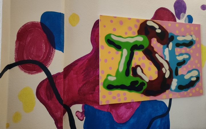 Eine bunte Wandmalerei zeigt abstrakte Formen in Rot, Blau und Gelb. Im Vordergrund hängt ein rechteckiges Bild mit gelbem Hintergrund und rosa Punkten. Darauf sind die Buchstaben 'ISE' in grüner, brauner und blauer Farbe mit plastischem Effekt gemalt.