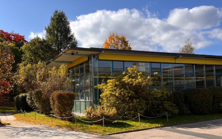 Ein moderenes Gebäude mit großen Glasflächen und flachen Dächern stehen in einer gepflegten Grünanlage. Der Himmel ist klar und blau.