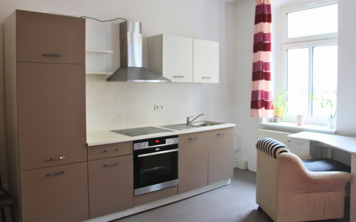 Küchenzeile mit hellbraunen und weißen Fronten, integriertem Backofen, Cerankochfeld und Dunstabzugshaube aus Edelstahl. Links ein hoher Schrank, rechts eine Spüle mit Unterschrank. An der Wand ein Fenster mit rot-weiß gestreiftem Vorhang, davor ein Schreibtisch mit einem Korbsessel. Auf dem Fensterbrett stehen mehrere grüne Pflanzen.
