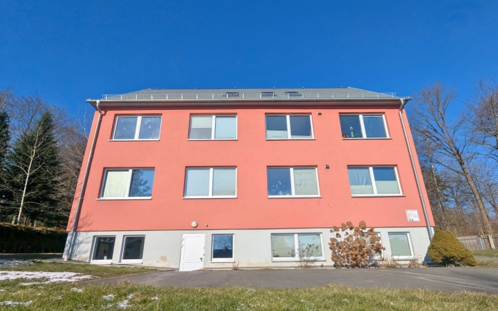 Das Foto zeigt ein großes rotes Haus mit acht Fenstern. Es ist frontal und von unten fotografiert. Im Hintergrund ist blauer Himmel und Wald. Im Vordergrund ist Wiese. 