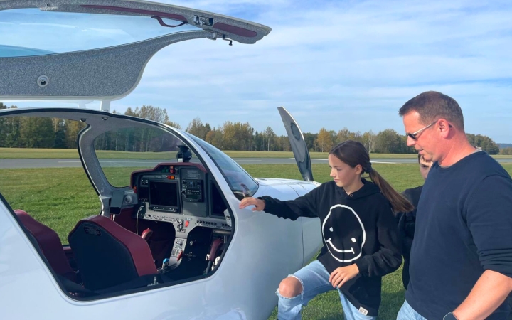 Zwei Personen stehen neben einem kleinen weißen Flugzeug auf einer Wiese. Die Cockpittür ist geöffnet, das Cockpit mit mehreren Anzeigen und Bedienelementen ist gut sichtbar. Eine Person trägt einen schwarzen Kapuzenpullover mit einem großen Smiley-Motiv, die andere ein dunkelblaues Oberteil. Beide blicken in das Cockpit.