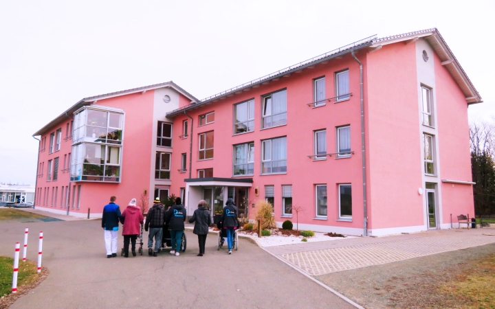 Im Hintergrund des Bildes sieht man ein großes rotes Haus, mit vielen Fenstern. Das Haus sieht einladend aus. Im Vordergrund sieht man eine Reihe aus Menschen, welche auf das Haus zugehen. Man sieht den Rücken der Personen. Einige von Ihnen tragen eine Di