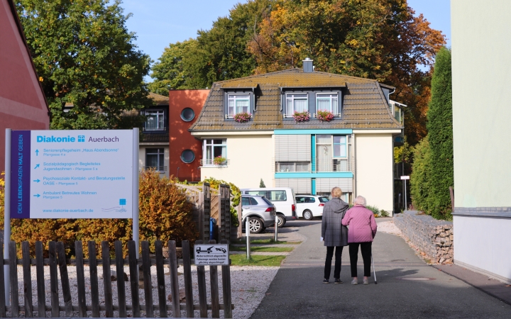 Auf dem Bild ist ein gelbes Haus zu sehen. Im Hintergrund ist ein herbstlicher Park. Im Vordergrund sieht man zwei Frauen auf das Haus zulaufen. Die eine Frau stützt die andere. Im Vordergrund links ist eine Infotafel zu sehen. Auf dieser sind verschieden