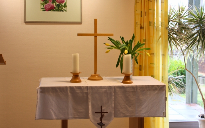 Ein kleiner Altar mit weißem Tuch, darauf ein Holzkreuz und zwei Kerzenständer mit weißen Kerzen. Rechts steht eine Vase mit gelben Blumen. Im Hintergrund hängen gelbe Vorhänge und ein Gemälde mit rosa Blumen an der Wand, rechts ist ein Fenster mit Blick nach draußen und einer Topfpflanze zu sehen.