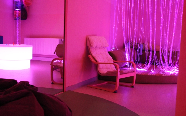 Ein gemütlicher, in rosa und lila Licht getauchter Raum, ausgestattet mit einem weißen Sessel und einer weichen Matte. Im Hintergrund hängen viele leuchtende Lichtfasern, die für eine entspannende Atmosphäre sorgen, während eine beleuchtete Blasensäule einen weiteren Sinnesanreiz bietet. Der Raum wirkt ruhig und lädt zum Wohlfühlen ein.