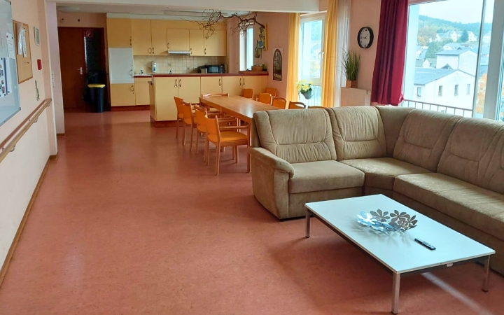 Ein Wohnraum für Wohn- und Esszimmer mit Küche. Im Hintergrund ist eine gelbe Küche, davor ein großer Esstisch. Im vordergrund ist ein Sofa mit Couchtisch. Es wirkt hell und einladend.