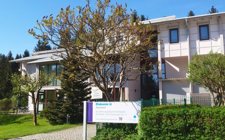 Zu sehen ist ein modernes, helles Gebäude mit großen Glasfronten und weißen Fassadenteilen. Im Vordergrund steht ein noch unbelaubter, aber kräftiger Baum und mehrere immergrüne Sträucher. Es ist ein gepflegter Rasen samt gepflastertem Weg angelegt. Im unteren Bereich des Bildes steht ein großes Schild mit der Aufschrift 
