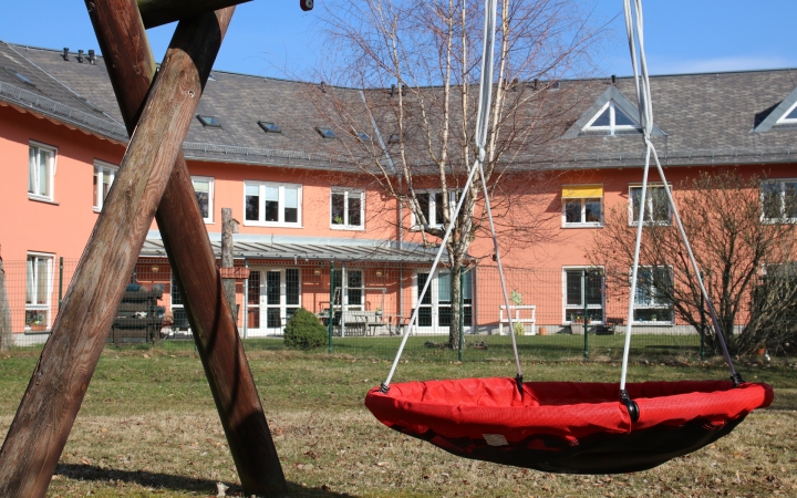 Im Vordergrund steht eine runde, rote Nestschaukel mit weißen Seilen, die an einem massiven Holzgestell befestigt ist. Im Hintergrund befindet sich ein zweistöckiges, lachsfarbenes Gebäude mit mehreren Fenstern und einem grauen Schrägdach. Der Garten ist von Rasen bedeckt, einige kahle Bäume stehen verteilt auf der Fläche. Die Szene ist von Tageslicht erhellt, der Himmel ist blau mit wenigen Wolken.
