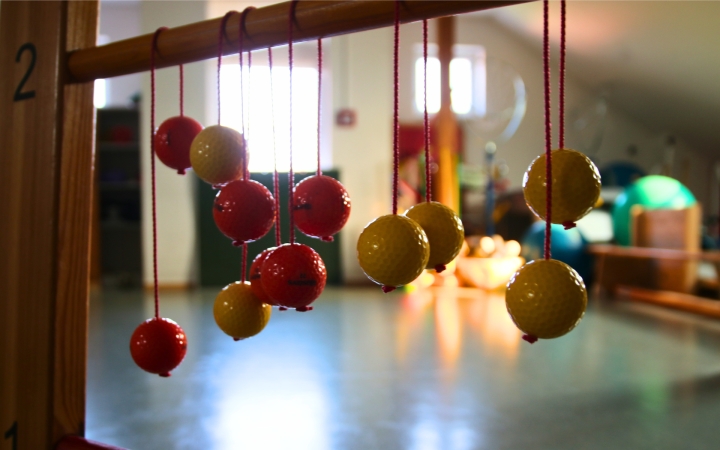 Im Vordergrund hängen mehrere gelbe und rote Kugeln an roten Schnüren von einer Holzstange. Die Kugeln sind glänzend und gleichmäßig rund. Im Hintergrund ist ein heller, unscharfer Raum mit Sportgeräten und Matten zu erkennen. Sonnenlicht fällt durch Fenster und erzeugt Lichtreflexe auf dem Boden.