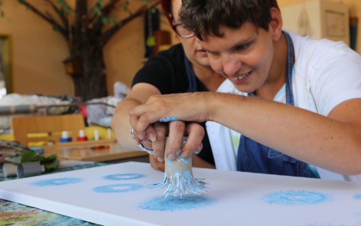 In einem Kreativraum sitzen zwei Personen an einem Tisch. Eine Person wird dabei von einer anderen angeleitet und unterstützt: Zusammen führen sie eine Hand, die mit blauer Farbe bestrichen ist, auf eine weiße Leinwand und stempeln mit den Fingern kreisförmige Muster. Im Hintergrund sind bunte Malutensilien wie Farben und Pinsel sowie Dekorationen und Möbelstücke zu erkennen. Die Szene vermittelt kreatives, gemeinschaftliches Arbeiten und künstlerische Förderung.