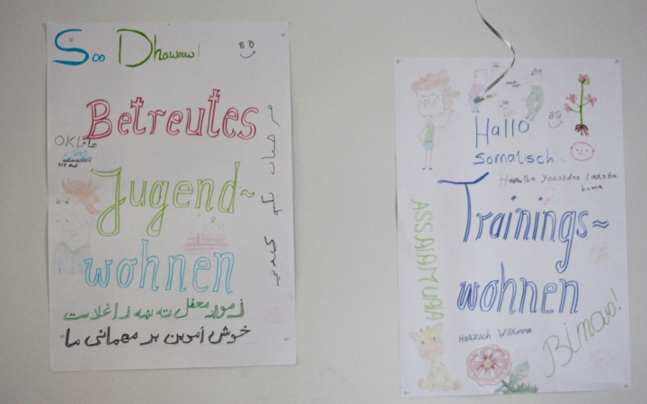 Zwei handgemalte Poster hängen an einer Wand. Auf dem linken steht in großen, bunten Buchstaben „Betreutes Jugendwohnen“ mit zusätzlichen kleinen Zeichnungen und Texten, teilweise auch in einer anderen Sprache. Auf dem rechten Poster steht „Trainingswohnen“, ebenfalls farbig gestaltet, mit Zeichnungen, Verzierungen und Willkommensbotschaften wie „Hallo“ und „Herzlich Willkommen“.
