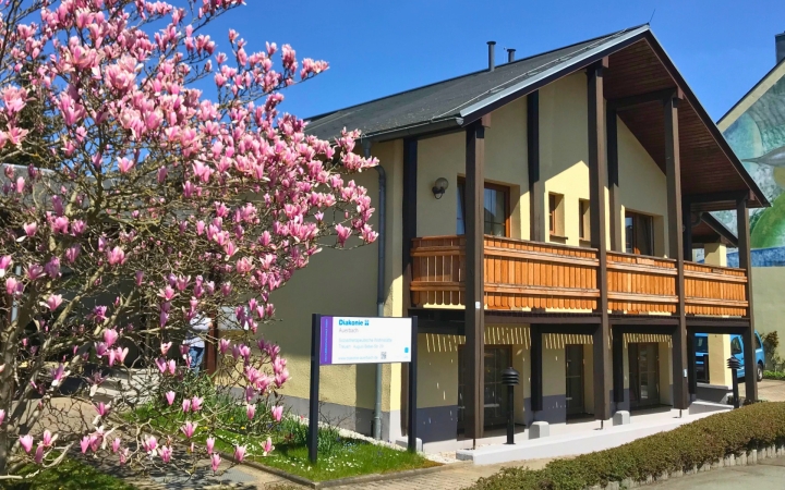 Ein freundliches, gelb gestrichenes Haus mit einem großen Vorbau aus dunklem Holz dominiert das Bild. Das Haus hat einen langen Balkon mit einem rustikalen Holzgeländer und mehrere Fenster. Links vorne steht ein prachtvoll blühender Magnolienbaum voller rosa Blüten, der einen Großteil des Vordergrunds einnimmt. Ein Schild mit dem Logo der Diakonie weist auf die Einrichtung im Haus hin. Das Wetter ist sonnig, der Himmel strahlend blau, die Atmosphäre wirkt einladend und frühlingshaft.