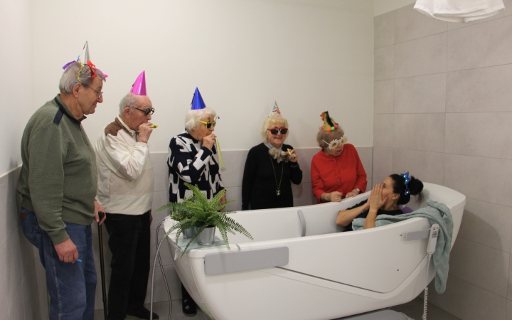 Auf dem Bild ist eine Badewanne zu sehen. In dieser sitzt eine Frau mit erschrockenem Gesichtsausdruck und hält sich die Hände vor den Mund. Um die Badewanne herum stehen drei Frauen und zwei Männer, welche auf die Badewanne schauen. Sie tragen alle einen
