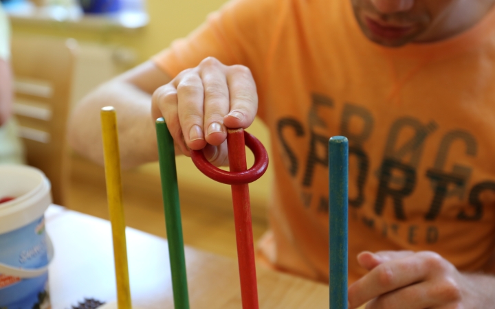 Nahaufnahme einer Person, die ein Motorikspiel spielt. Die Person trägt ein orangefarbenes T-Shirt und steckt einen roten Ring auf einen von mehreren bunten vertikalen Stäben (gelb, grün, blau, rot), die auf einem Holzbrett montiert sind. Im Hintergrund stehen weitere Spielmaterialien, wie ein Behälter mit weiteren Ringen. Die konzentrierte Haltung und der gezielte Griff zeigen eine Förderung der Feinmotorik und kognitiven Fähigkeiten, oft verwendet in therapeutischen oder pädagogischen Einrichtungen.
