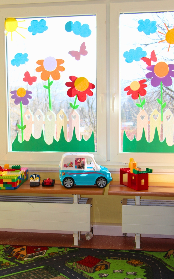 Bunte Papierblumen, Wolken, Schmetterlinge und eine weiße Zaunreihe sind an zwei großen Fenstern angebracht. Auf den Fensterbänken stehen Bauklötze, ein Spielzeugauto und weitere Spielzeuge. Im unteren Bildbereich ist ein Teppich mit Straßenmotiven zu sehen.