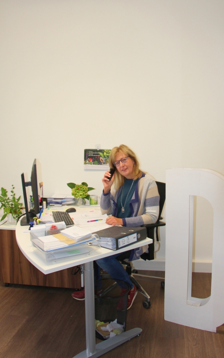 Eine Person sitzt an einem weißen Schreibtisch und telefoniert. Auf dem Tisch liegen mehrere Aktenordner, Papierstapel und Schreibutensilien. Zwei Monitore stehen nebeneinander, dahinter sind grüne Zimmerpflanzen platziert. Die Person trägt eine helle Strickjacke und rote Schuhe. Der Raum wirkt hell und aufgeräumt, die Wand ist weiß. Im Hintergrund hängt ein kleines Poster mit Pflanzenmotiv.
