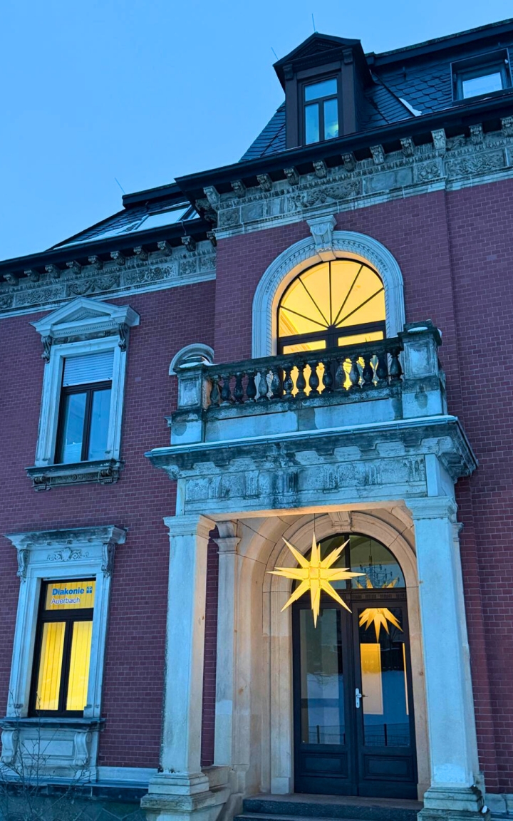 Das Bild zeigt den Eingang eines Backsteingebäudes. Der Eingang ist mit zwei Säulen umrandet. Im Eingang hängt ein großer leuchtender Stern. Neben dem Eingang links ist ein fenster mit der Schrift 